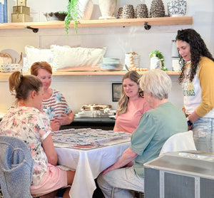A group of five women gathered around a table in a bright, modern home decor showroom learning how to play American Mahjong. Shelves in the background display ceramic vases, pillows, and decorative items.