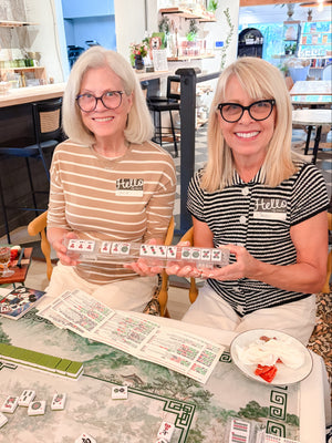 Two smiling women sitting at a table playing a mahjong game in a bright indoor shop or cafe. One woman wears a beige and white striped shirt with 'Hello' printed on it, and the other wears a black and white striped shirt.